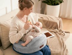 New mom smiling while breastfeeding baby on gray Momcozy Original nursing pillow in sunny living room