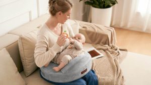 New mom smiling while breastfeeding baby on gray Momcozy Original nursing pillow in sunny living room