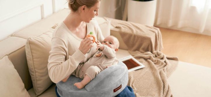 New mom smiling while breastfeeding baby on gray Momcozy Original nursing pillow in sunny living room