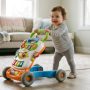 Smiling baby pushing a colorful wooden activity walker across a bright living room.