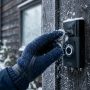 Photorealistic close-up of a person's gloved hand gently brushing frost and snow away from a modern video doorbell mounted on a dark wooden exterior wall on a chilly winter morning, soft natural daylight, shallow depth of field, commercial lifestyle product photography.